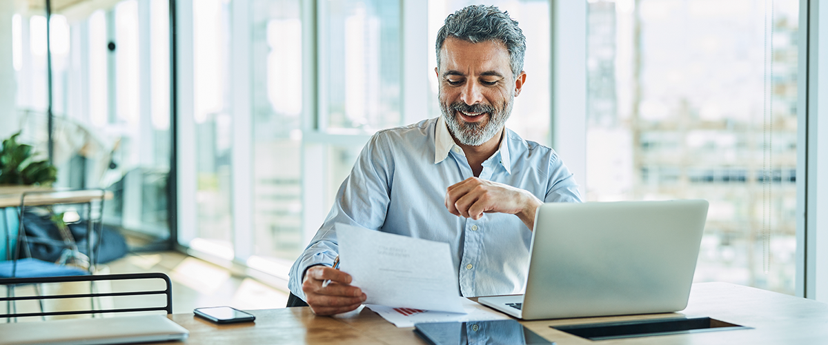 Man in office setting reading off of a sheet of paper, sitting in front of a laptop.