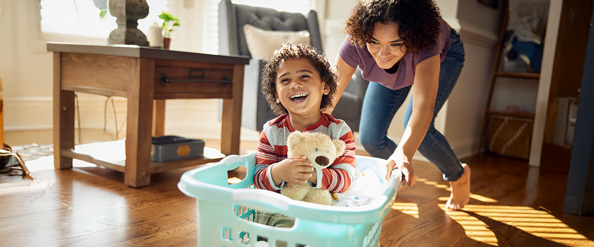 A young boy holding stuffed bear, being pushed around in a laundry basket.