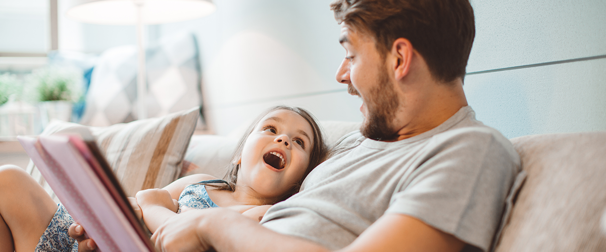 Father and a young daughter, sitting on the couch together reading a book.